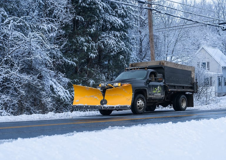Black snow plow truck with yellow blade removing snow from a residential street lined with snow-covered trees and houses.