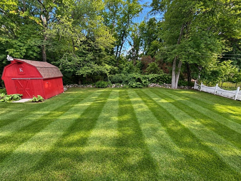 Red barn on manicured lawn with alternating stripe pattern, surrounded by mature trees and white fence