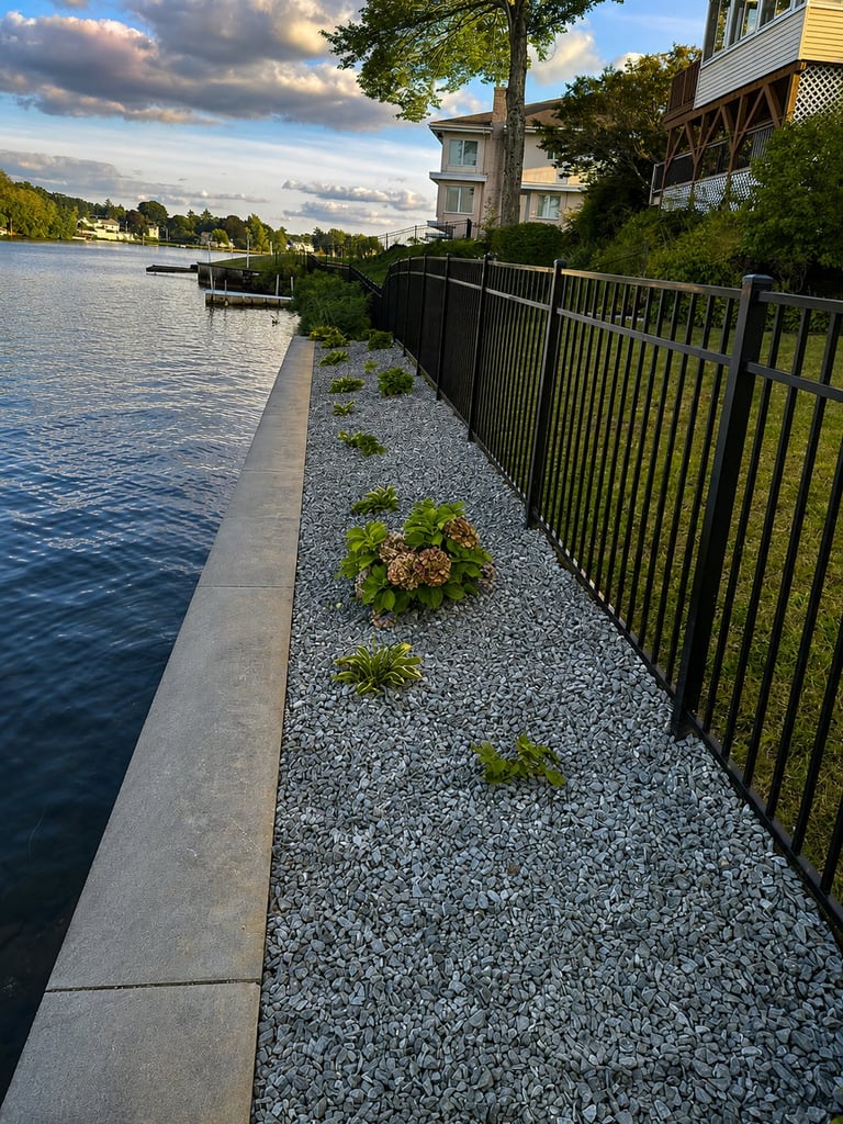 Waterfront property with gravel strip, black metal railing, and modern house overlooking a calm river under cloudy sky.